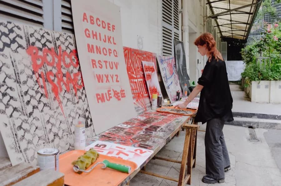 Atelier en extérieur : alphabets et slogans peints en rouge sur des panneaux, une personne travaille à une table.