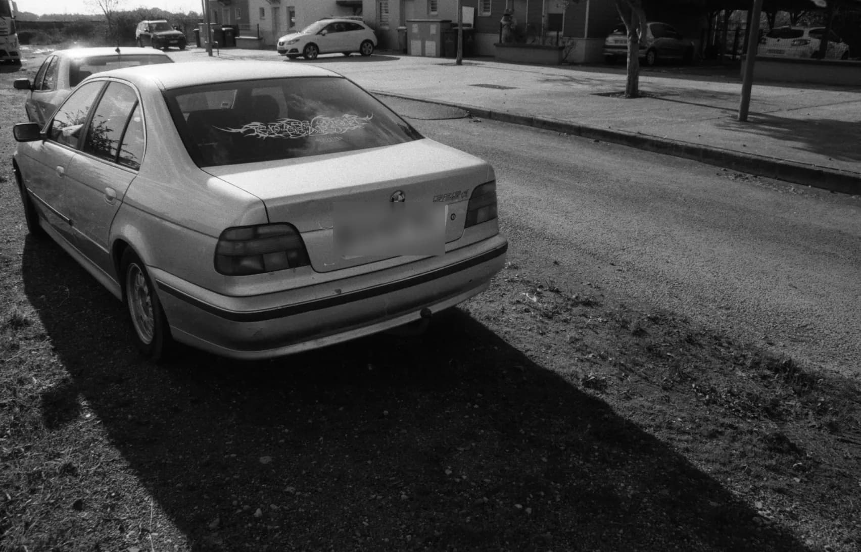 A saloon car parked at the edge of a residential street, licence plate blurred, black and white