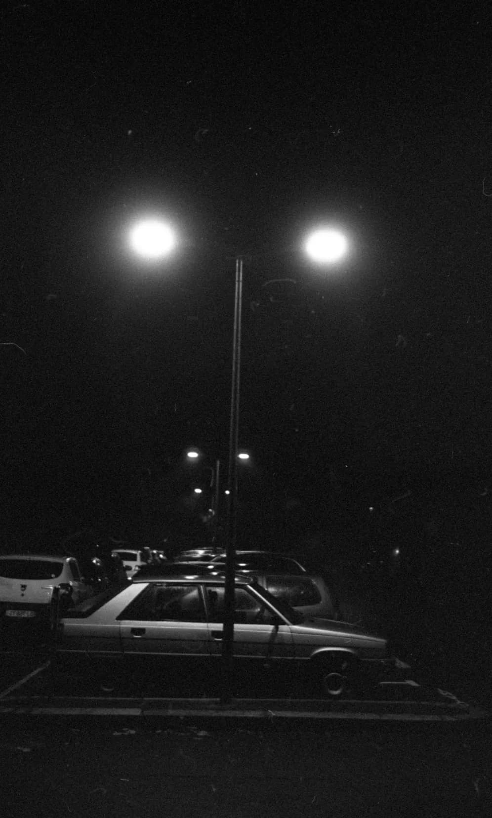 A double streetlamp lit above a car park at night, black and white
